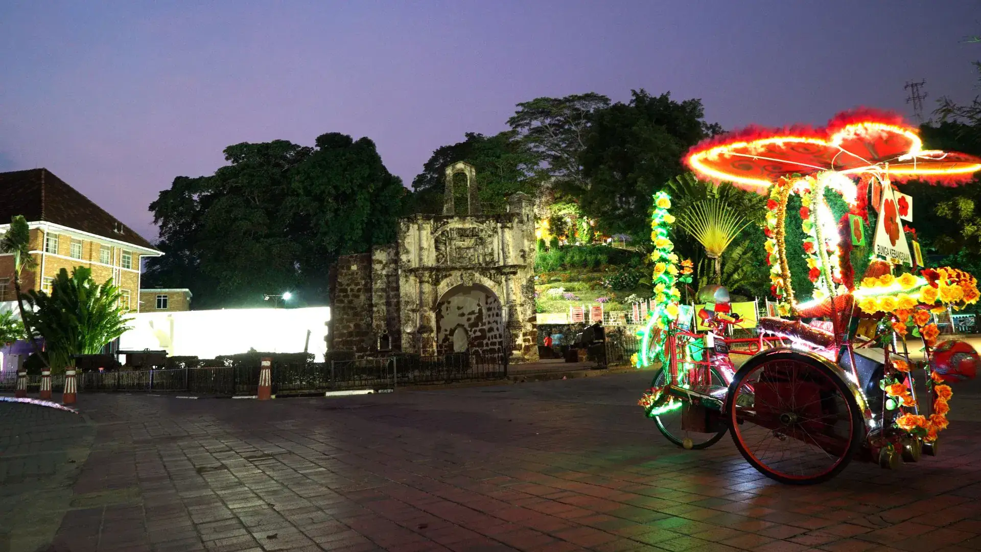 A trishaw strung with lights and flowers parked at night outside the A Famosa gate