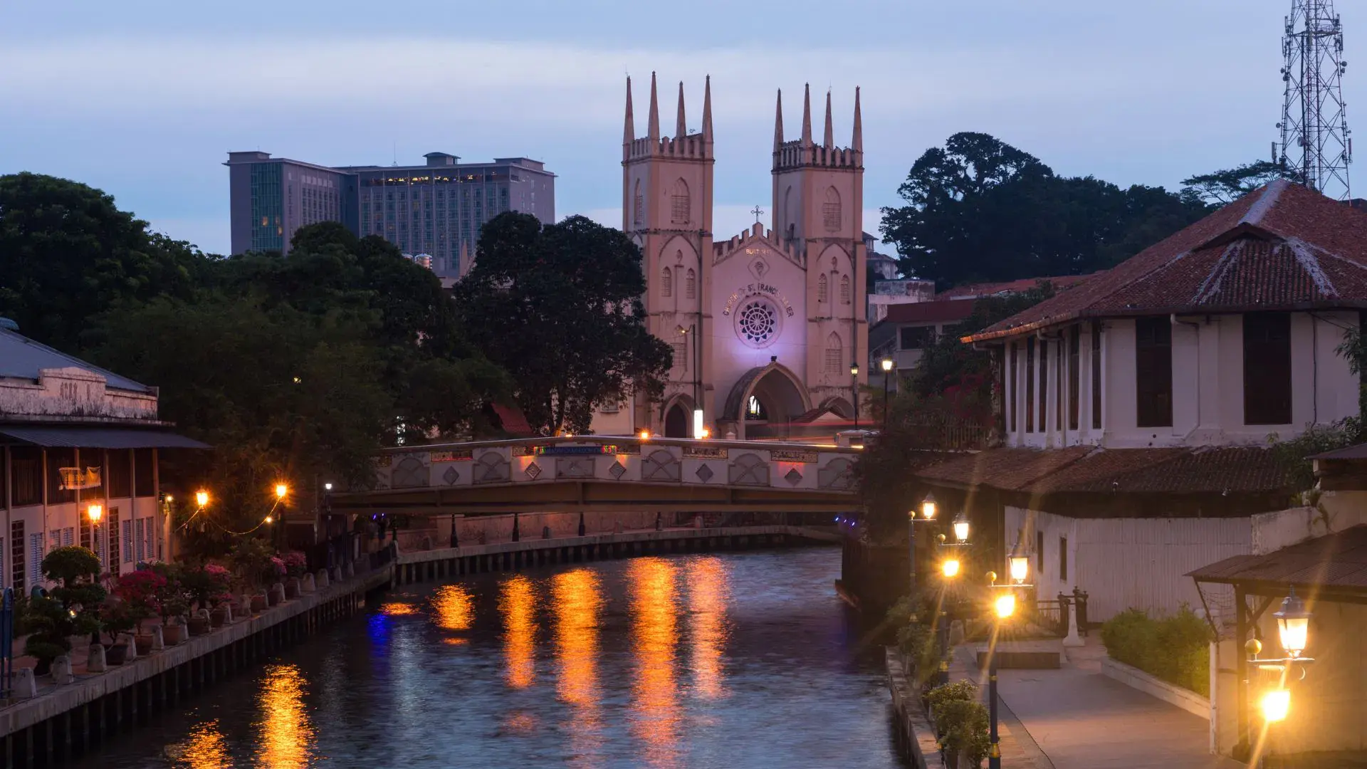 The Melaka River at dusk, lamps lit along both banks and the twin spires of the Church of St Francis Xavier behind the bridge