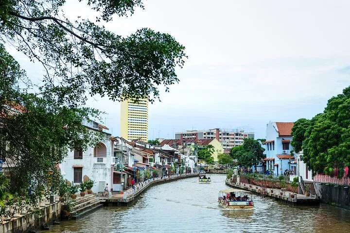 A river cruise boat on the Melaka River in daylight, passing riverside shophouses