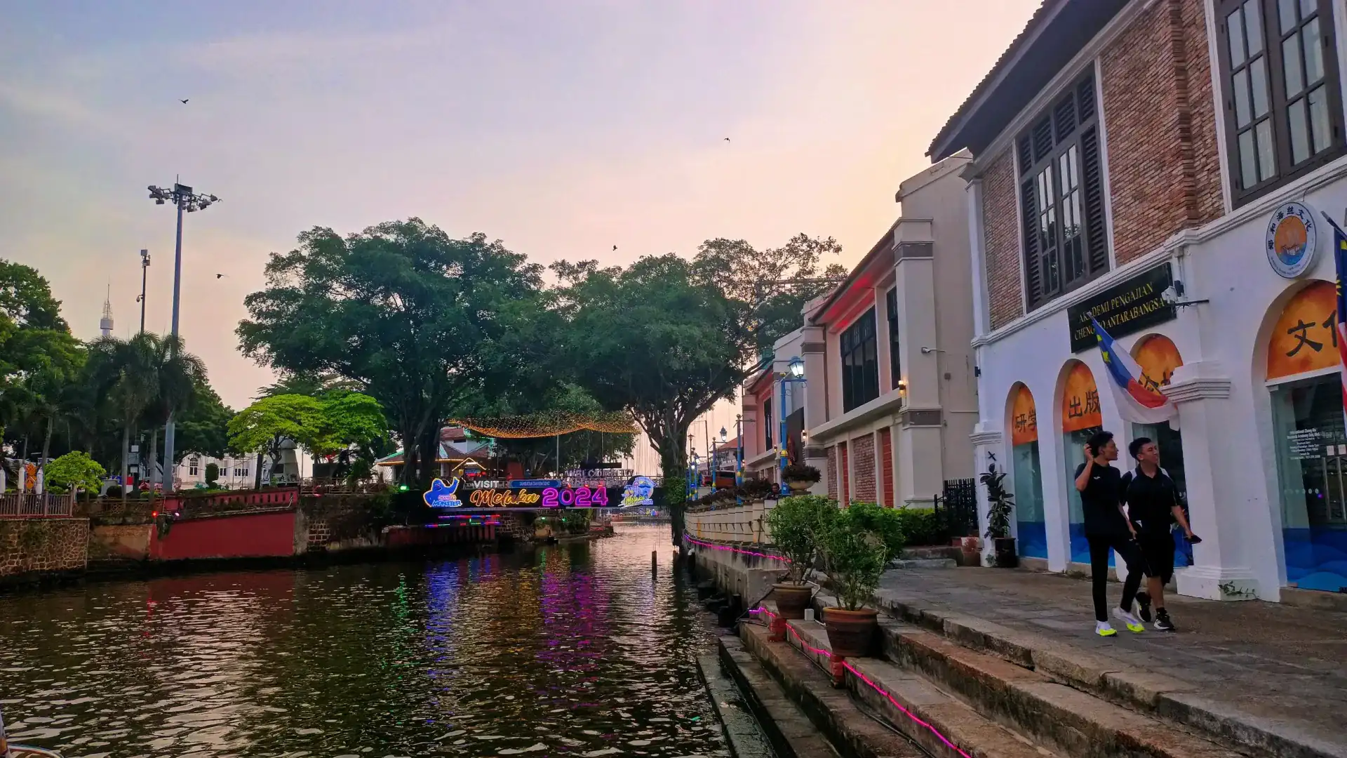 The Melaka riverwalk at dusk, with riverside shophouses along the far bank