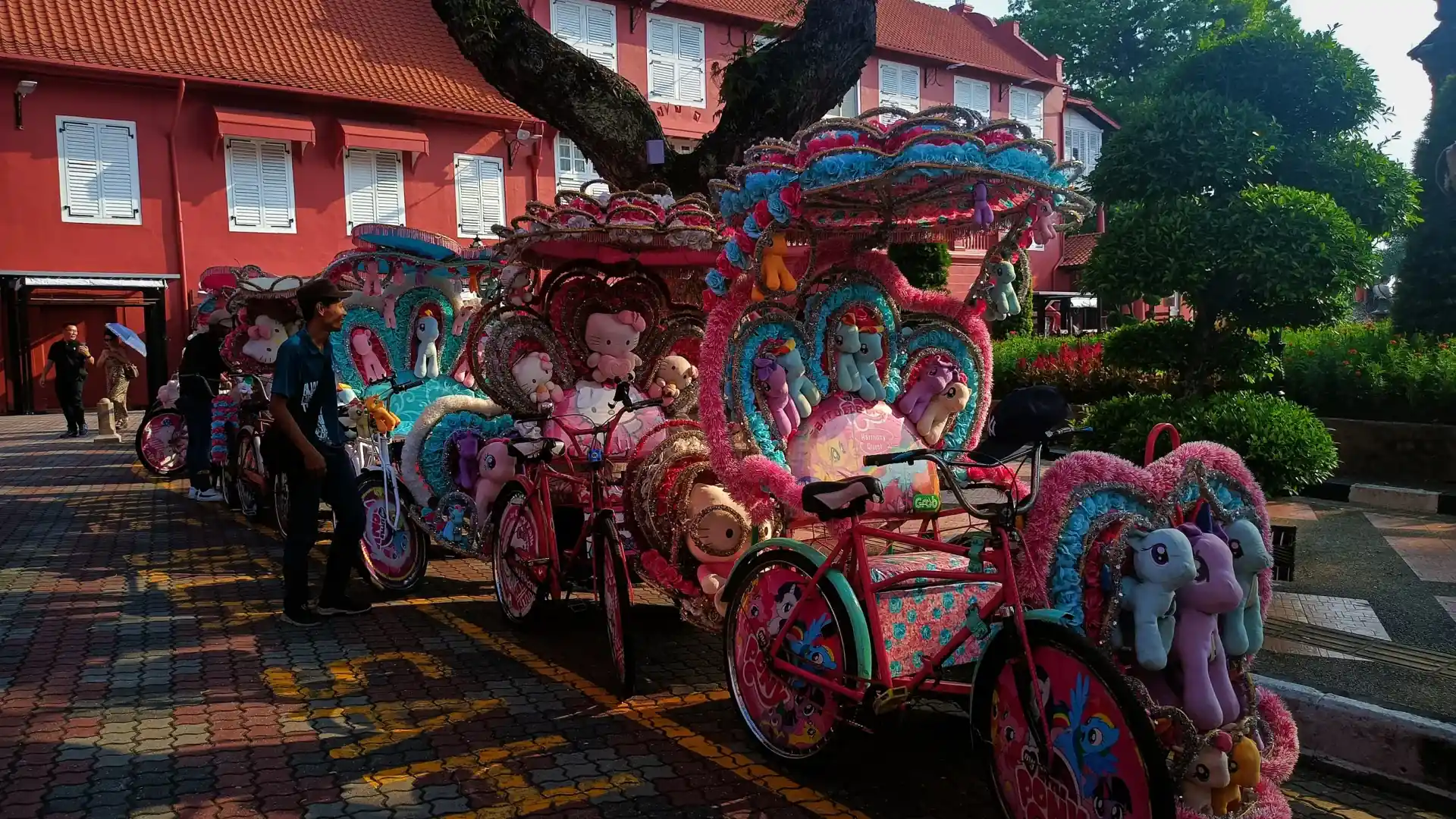 A row of decorated trishaws parked outside the red Stadthuys buildings, a rider standing beside them